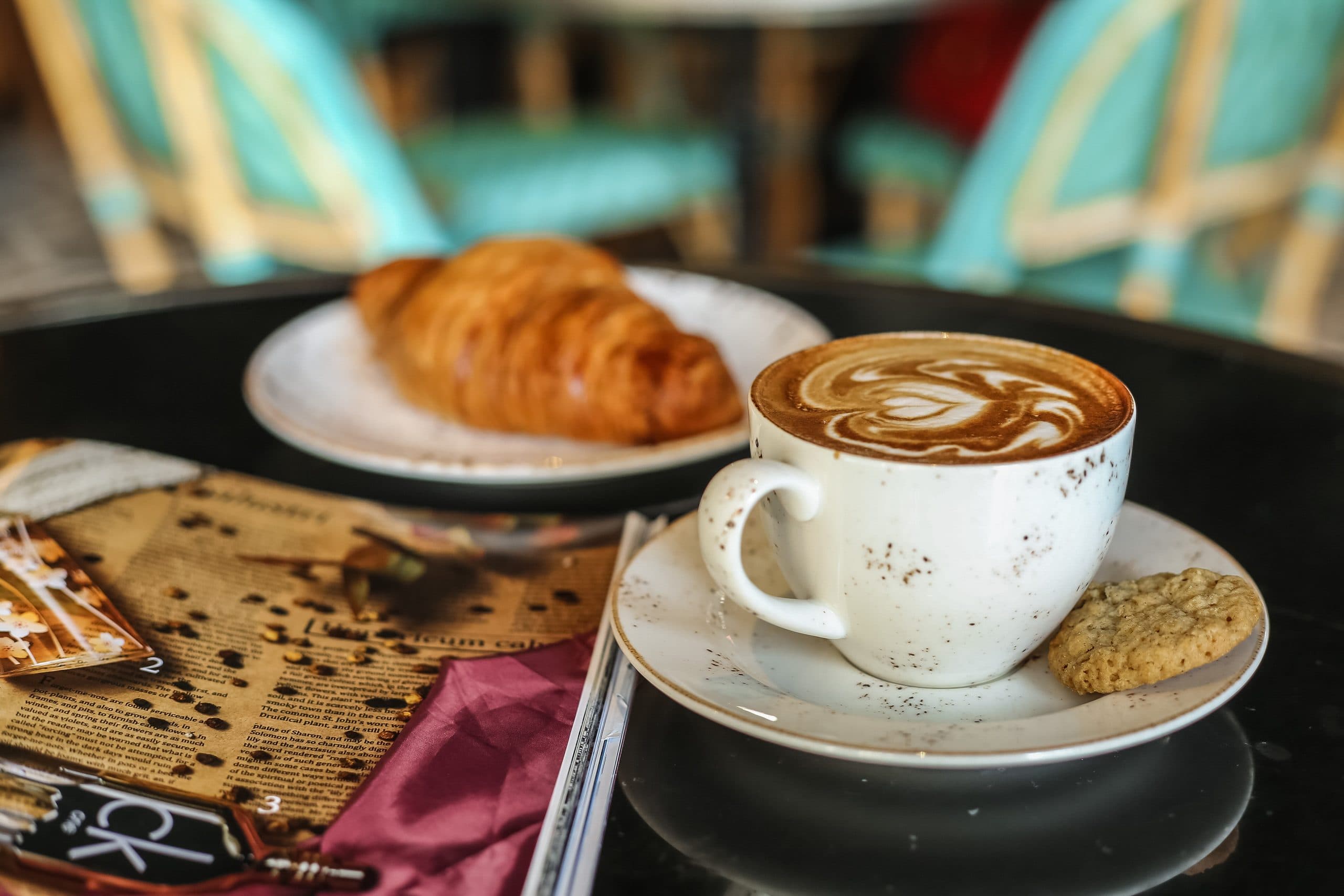 Cappuccino mit Latte Art, daneben ein Keks und ein Croissant auf einem dunklen Tisch
